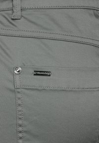 Närbild av en grå tygbyxans ficka med synlig sömnad, metallnitar och en liten rektangulär varumärkesetikett.