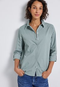 Vrouw met krullend haar die een groen en wit verticaal gestreept gebuttond shirt en blauwe spijkerbroek draagt, met handen in de zakken, staand tegen een lichte muur.