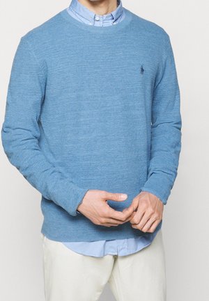 Man draagt een lichtblauwe trui met ronde hals over een blauw overhemd met knopen en witte broek, handpalmen voor de taille samengevouwen.