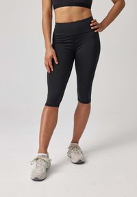 Zwarte capri leggings met een hoge tailleband, een gladde textuur en duidelijke naden, gecombineerd met lichtgekleurde sportschoenen.