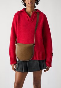 Brązowa skórzana torebka crossbody o zaokrąglonym kształcie, z perforowanym detalem logo i regulowanym paskiem. Noszona na czerwonym swetrze z dzianiny i w kratkowanej spódnicy.