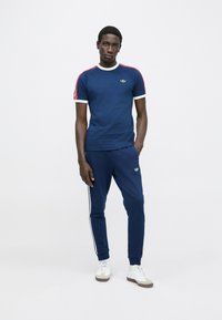 Tamsiai mėlynas medvilninis marškinėlis su baltomis ir raudonomis dryžuotomis rankovėmis, apvalia iškirpte ir auksiniu logotipu. Derinamas su atitinkančios spalvos mėlynais sportinėmis kelnėmis, turinčiomis baltas šonines juostas.