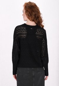 Pull en tricot noir à coupe ample, présentant un motif de tissage ouvert et des poignets côtelés. Le dos affiche un design texturé et un détail de marque.