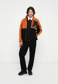 Chaqueta de forro polar naranja y negra con cremallera, que presenta un bolsillo en el pecho y un material texturizado. Lleva puesta con pantalones y zapatos negros.