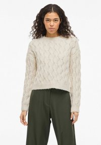 Pull beige clair en maille avec un motif torsadé texturé. Col rond et poignets côtelés. Porté avec un pantalon vert foncé.