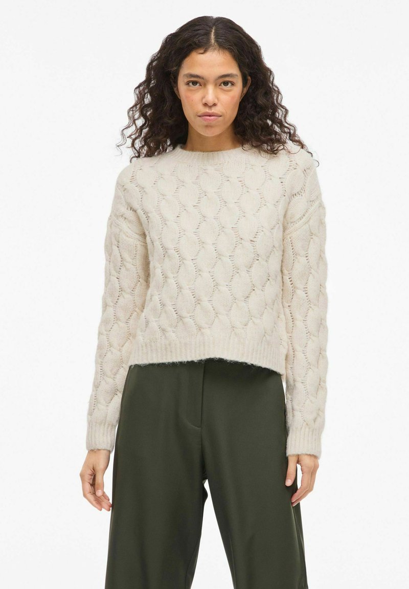 Pull beige clair en maille avec un motif torsadé texturé. Col rond et poignets côtelés. Porté avec un pantalon vert foncé.