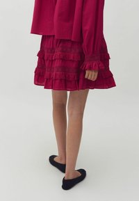 Rochie roșie stratificată cu tivul volanat și accente brodate, asociată cu pantofi negri cu talpă joasă. Țesătură moale, ușoară, cu un design fluid.