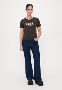 Ruda trumpa rankovė t-shirt su balta užrašu "MUSE Energy", derinama su mėlynomis plačiomis džinsomis ir juodais platforminiais batais, priešais baltą foną.