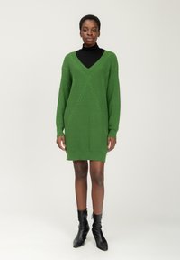Abito maglione verde a collo a V, con motivi a trecce diagonali e maniche lunghe. Indossato con un dolcevita nero e stivali alla caviglia.