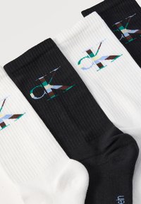 Τέσσερις ραβδωτές κάλτσες σε εναλλασσόμενα μαύρο και λευκό, με κάθε μία να διαθέτει πολύχρωμο λογότυπο "CK" κοντά στην κορυφαία μανσέτα.