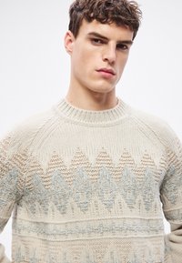 Jasnobeżowy sweter z dzianiny z geometrycznym wzorem w stonowanym niebieskim i kremowym kolorze. Prążkowany, okrągły dekolt oraz teksturowane akcenty na rękawach.