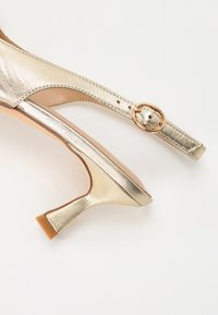 Zapato tipo slingback metálico dorado con un pequeño tacón, que presenta un acento de hebilla redonda y una textura suave. Correa ajustable para un mejor ajuste.
