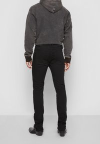 Black fitted jeans with a smooth texture, paired with a dark, distressed hoodie, visible stitching, and slim-fit design. Black ankle boots complete the look.