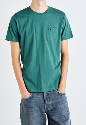 Mann trägt grünes kurzärmeliges T-Shirt mit Brusttasche und schwarzem Label, kombiniert mit weiten blauen Jeans, Hände in den Taschen.