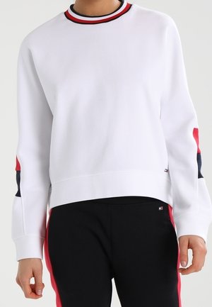 Persoon draagt een witte trui met rode en marineblauwe strepen op de kraag en mouwen, gecombineerd met een zwarte broek met rode zijstrepen.