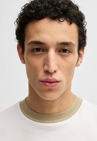 Maglietta bianca con colletto a coste beige. Il modello ha i capelli corti e ricci e una pelle liscia, mostrando toni neutri e caratteristiche naturali.