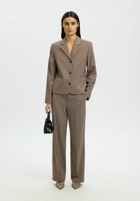Costume marron sur mesure avec une veste cintrée à simple boutonnage, un pantalon large assorti et des talons beiges pointus. Sac à main noir en accessoire.