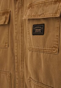 Tan fabric jacket front with stitched flap pockets, featuring a black label reading "ORIGINALS GUESS Authentic Style Designed in Los Angeles."