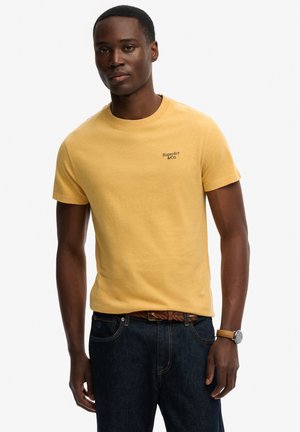 Mand iført en sennepsgul t-shirt med korte ærmer og "Superdry & Co"-logo, mørkeblå jeans, brunt flettet bælte og brun ur på venstre håndled.