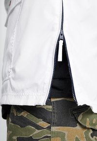 Biała kurtka na zamek błyskawiczny z granatowymi akcentami, z widocznym zamkiem i bocznymi kieszeniami, w połączeniu z spodniami w kamuflaż.