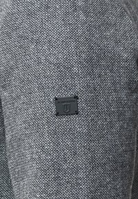 Tissu gris texturé avec un petit détail d'étiquette noire Jean's Code. Le motif de tissage est serré et uniforme, offrant une apparence durable.