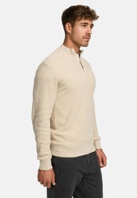Beige kvart-lynlås pullover trøje med ribbet tekstur, lange ærmer og lige kant. Parret med mørkegrå bukser.