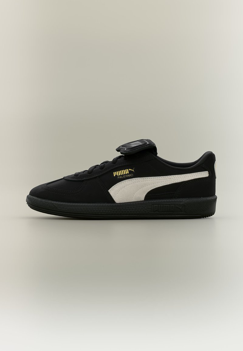 Sneaker Puma Palermo nera con striscia bianca, logo dorato, suola nera e coprilacci su sfondo grigio chiaro.