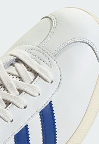 Bijela kožna tenisica s teksturiranom površinom, plavim akcentima u obliku cik-cak pruga, ravnim bijelim vezicama i kremastom gumiranom potplatom.