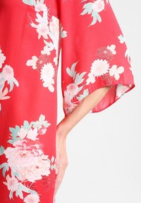 Kimono floral rouge avec des manches larges, présentant de grandes fleurs rose clair et teal. Tissu doux et lisse avec un design fluide.