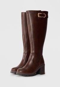 Bottes en cuir marron jusqu'au genou avec un talon épais, présentant une boucle en or et de subtils détails de couture sur le dessus.
