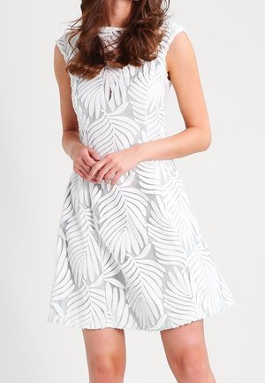 Biała sukienka bez rękawów z dużymi teksturowanymi wzorami liści, noszona przez osobę stojącą z rękami złożonymi z przodu.