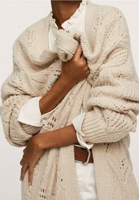 Beżowy sweter rozpinany na guziki z teksturowanym wzorem, noszony na białej koszuli z falbankami. Charakteryzuje się wygodnym, oversize'owym krojem z otwartym przodem.