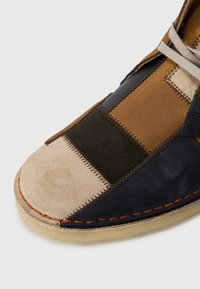 Patchwork shoe featuring brown, green, beige, and navy suede and leather. Notable hand stitching and a rubber sole with orange accents.