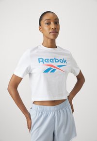 Balta trumpa marškinėliai su mėlynu ir raudonu "Reebok" logotipu. Trumpomis rankovėmis, apvaliu iškirpimu. Derinama su šviesiai mėlynomis šortais. Paprastas dizainas.