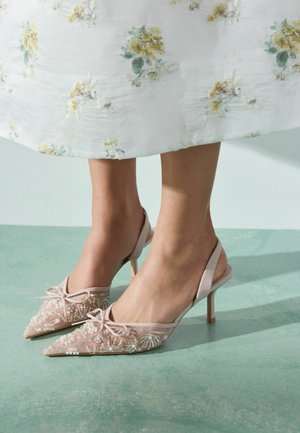 Kvinde iført lyserøde paliet-pointedtoe slingback hæle kombineret med en hvid blomster-midi-kjole, stående på grønt gulv.
