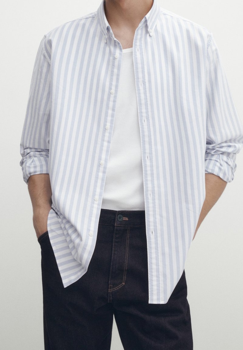 Homme portant une chemise à rayures verticales blanches et bleu clair par-dessus un débardeur blanc avec un jean bleu foncé, mains dans les poches.