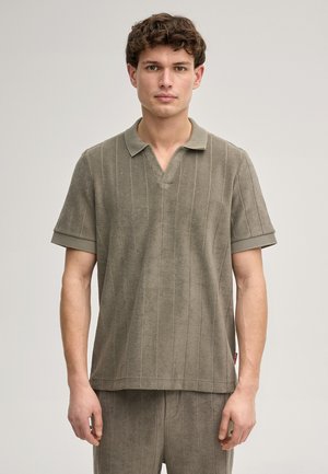 Jonge man met kort krullend haar, gekleed in een taupekleurig polohemd met korte mouwen en bijpassende broek, tegen een eenvoudige lichte achtergrond.