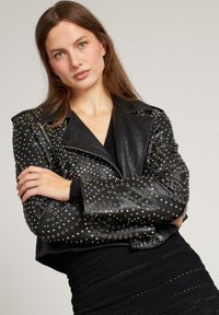 Giubbotto da motociclista in pelle nera adornato con borchie argentate, caratterizzato da un ampio colletto, tasche con zip e un design accorciato. Texture liscia.