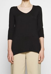 Svart långärmad tunika med V-ringning, tillverkad av mjuk tyg. Har en avslappnad passform och en något längre bakre nederkant. Matchas med beiga byxor.