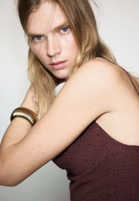 Bordový top bez rukávov s textúrovaným vzorom z malých korálikov; model má na zápästí zlatý kovový náramok. Svetlé pozadie.