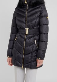 Schwarze Steppjacke mit Chevron-Steppmuster, goldenen Beschlägen, tailliertem Gürtel und Kapuze mit Kunstfell. Verfügt über seitliche Reißverschlusstaschen und Knopfmanschetten.