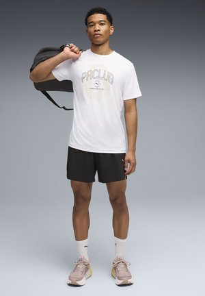 White t-shirt with "PRCLUB" graphic, black shorts, beige sneakers, and white socks; holding a black duffel bag over one shoulder.