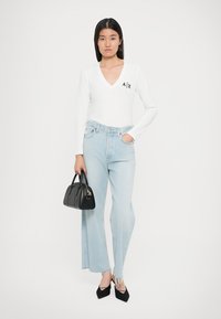 Top bianco a maniche lunghe con scollo a V e logo nero, jeans blu chiaro a vita alta con orli sfrangiati, borsa nera, tacchi a punta neri.