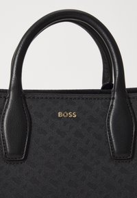 Fekete kézitáska texturált felülettel, bőr fogantyúkkal és arany "BOSS" logóval. Bonyolult mintázatokkal és sima, fényes részletekkel rendelkezik.