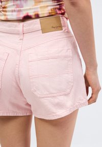 Γυναίκα που φοράει ανοιχτό ροζ τζιν σορτς με πίσω τσέπες και μία ταμπέλα από ταμπά δέρμα της Pepe Jeans London, σε συνδυασμό με ένα πολύχρωμο τοπ.