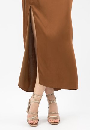 Jambes croisées portant des sandales à talons hauts métalliques à lanières et une jupe midi marron avec une fente latérale sur fond blanc.