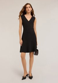 Rochie neagră fără mâneci, cu decolteu în V, corset ajustat, fustă evazată și textură fină. Poartă încălțări negre cu vârf ascuțit și o geantă mică.