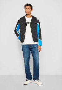 Veste matelassée noire avec des accents bleu et blanc, associée à un t-shirt blanc et un jean en denim bleu. Des baskets blanches complètent le look.