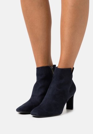 Bottes en daim bleu marine montant à la cheville, avec bouts pointus, fermetures éclair latérales et talons blocs, portées sur des jambes nues sur un fond clair.