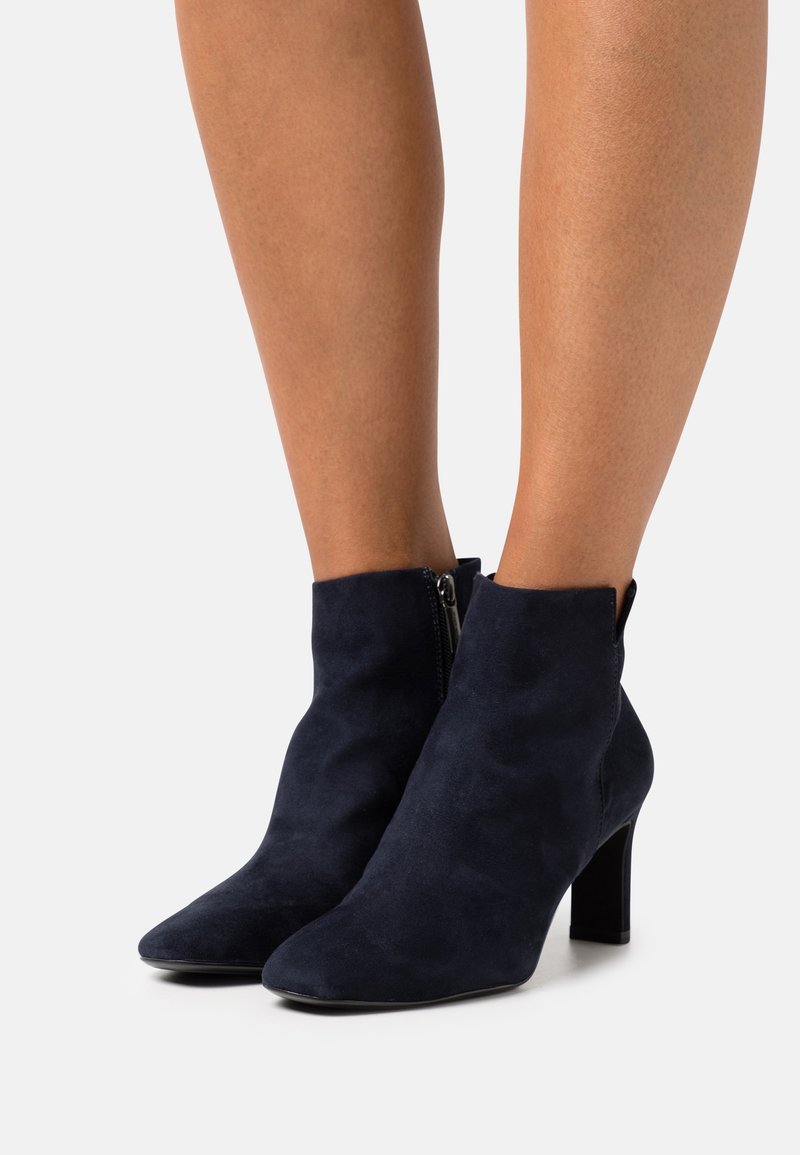 Bottes en daim bleu marine montant à la cheville, avec bouts pointus, fermetures éclair latérales et talons blocs, portées sur des jambes nues sur un fond clair.